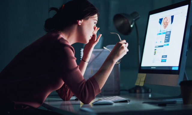 Woman in a dark room in front of a computer monitor, taking off her glasses and rubbing her eyes.