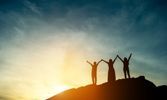 3 women in silhouette on a mountaintop with joined, raised hands