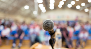 OCT 29 | Webinar: AOA Virtual Town Hall on Reimbursement and Coverage Fairness Close-up of a microphone on a podium with a blurry crowd of people in the background.