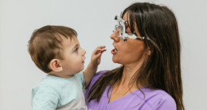 DEC 5 | Webinar: Visual Development: What Is Normal and What Is Not Woman optometrist holding a toddler boy in her arms while performing an eye exam using a phoropter.