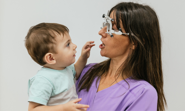 Woman optometrist holding a toddler boy in her arms while performing an eye exam using a phoropter.