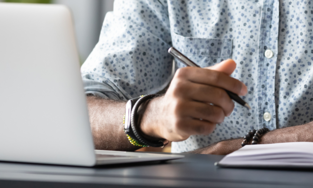 Close up of a man taking notes in front of a laptop.