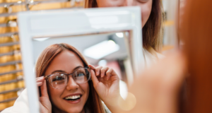 5 Proven Optical Promotions To Boost Your Capture Rate a woman trying on glasses, looking in a mirror at an optical