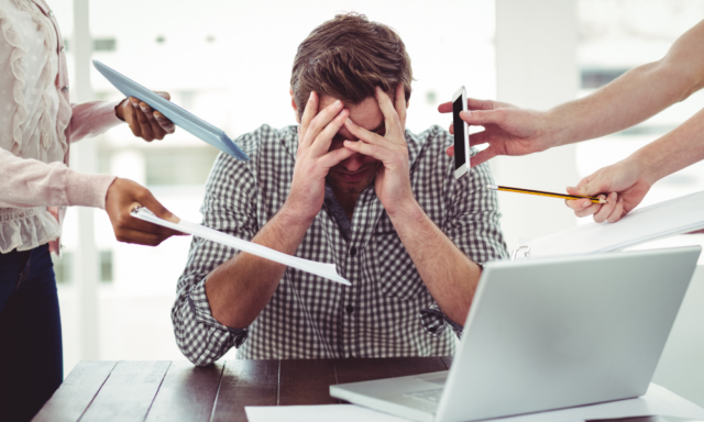 Man at a laptop with his head in his hands, surrounded by people who want his attention